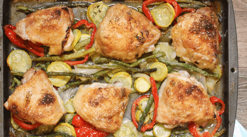 A sheet pan with six roasted chicken thighs laying on a bed of yellow squash and asparagus.